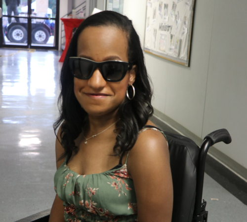 Young woman in a floral dress and sunglasses smiles confidently while seated in a wheelchair.